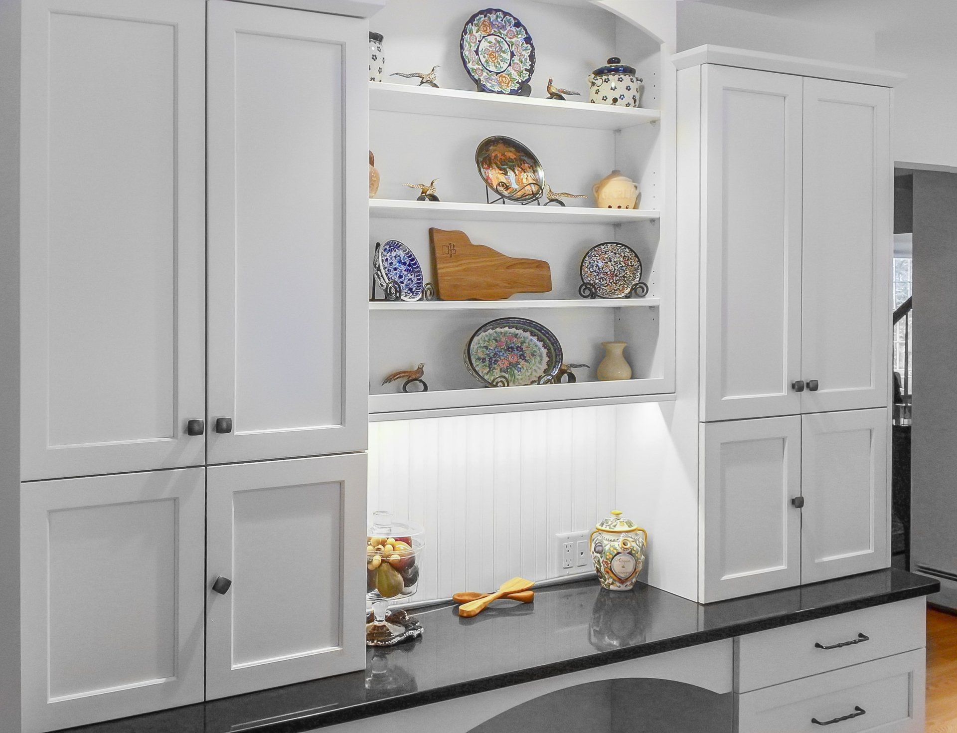 White kitchen cabinets with built-in open shelves displaying plates and decor above a dark countertop.