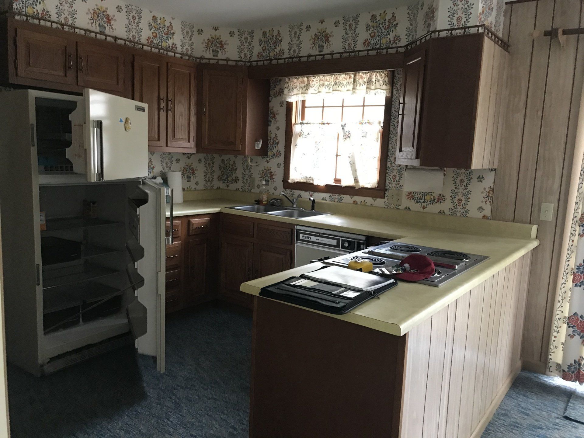 Small kitchen with wood cabinets, white appliances, patterned wallpaper, and a window over the sink.