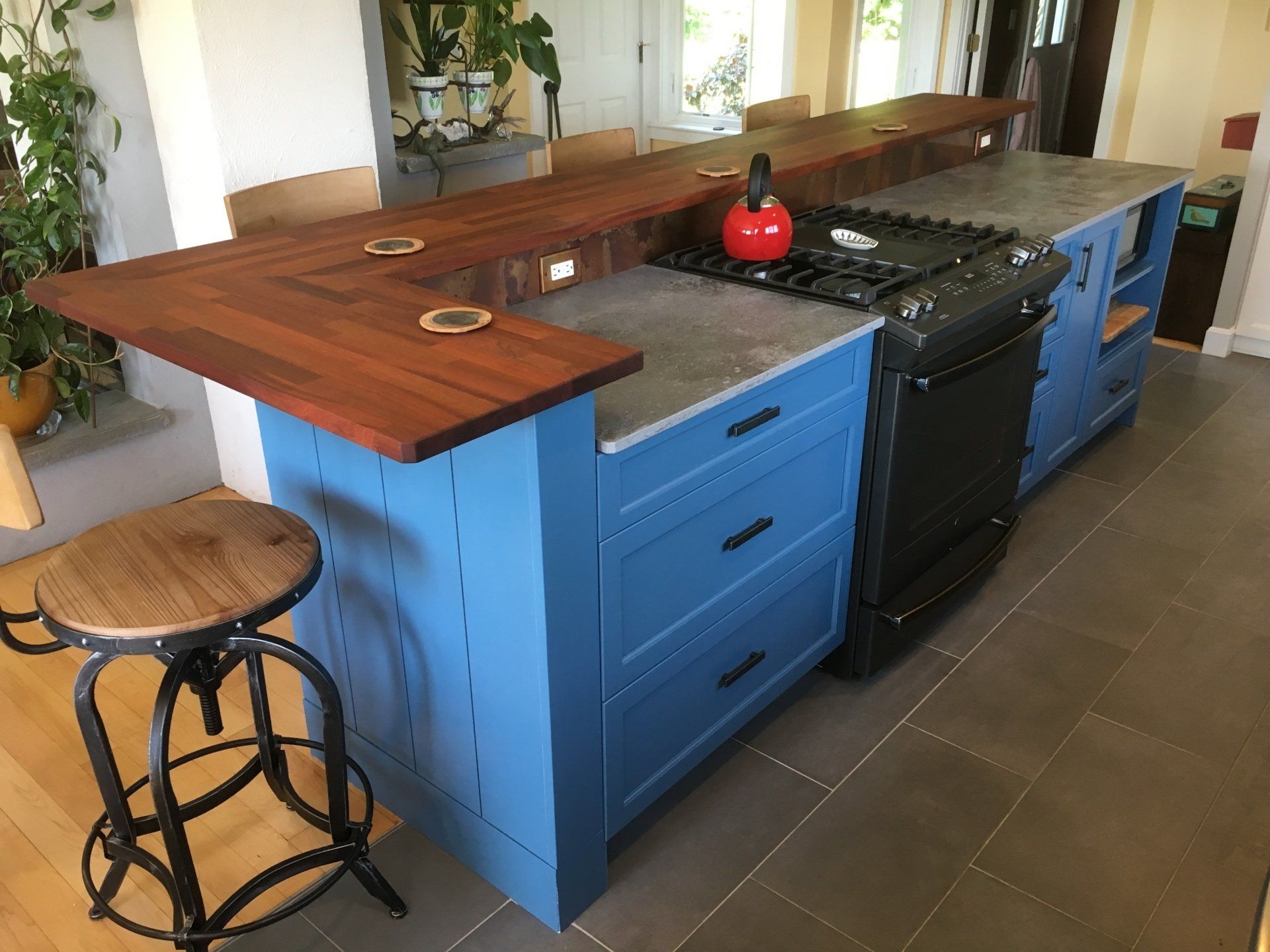 Blue kitchen island with wooden countertop, black stove, and a round stool in a bright room