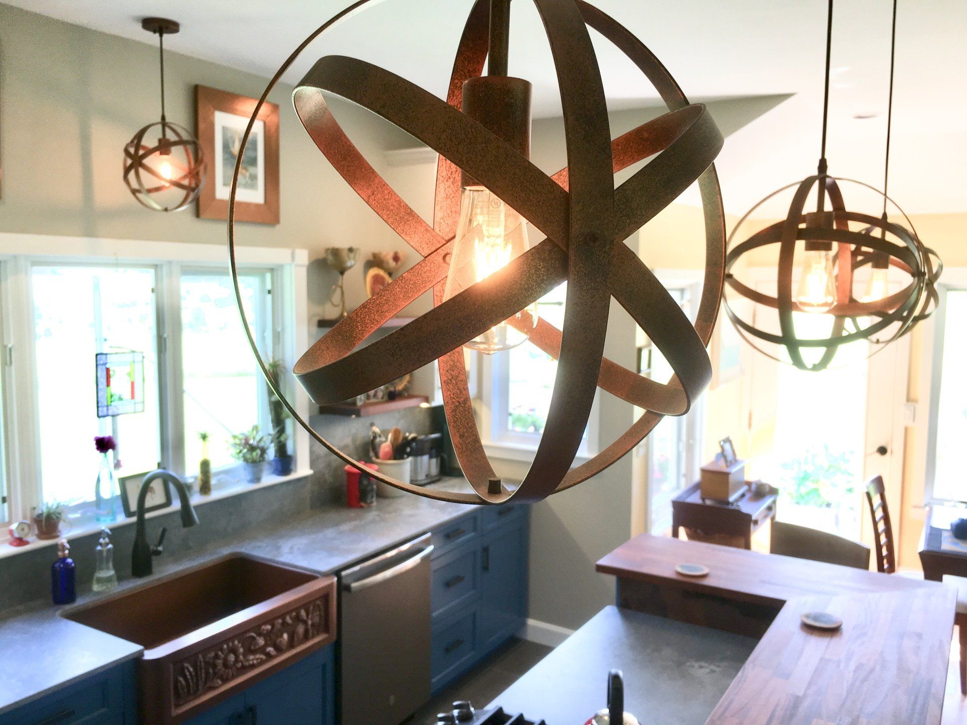 Modern kitchen with hanging orb lights, blue cabinets, and a large abstract metal wall sculpture.
