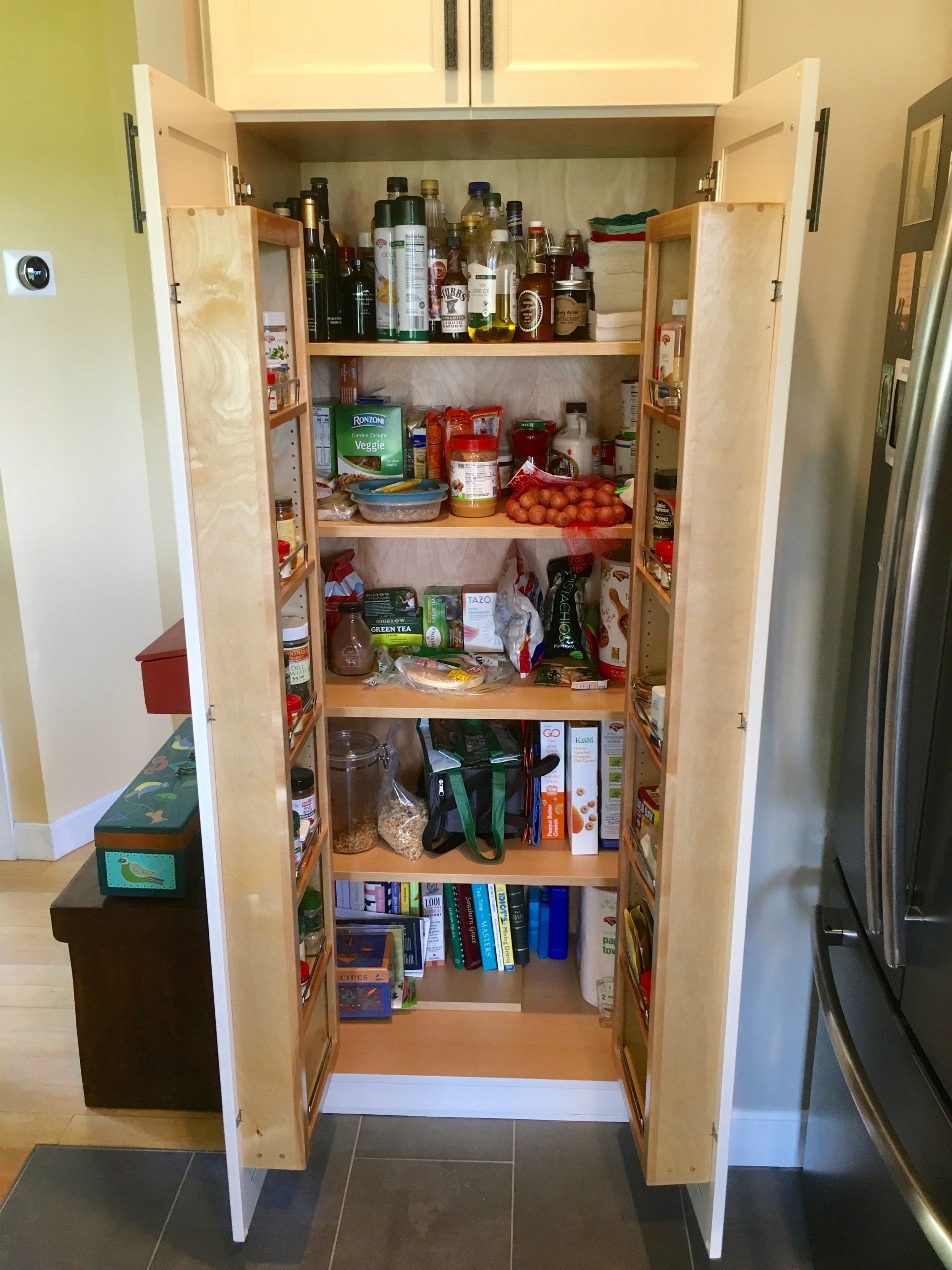 Open pantry with shelves of jars, cans, and snacks beside a stainless steel refrigerator.