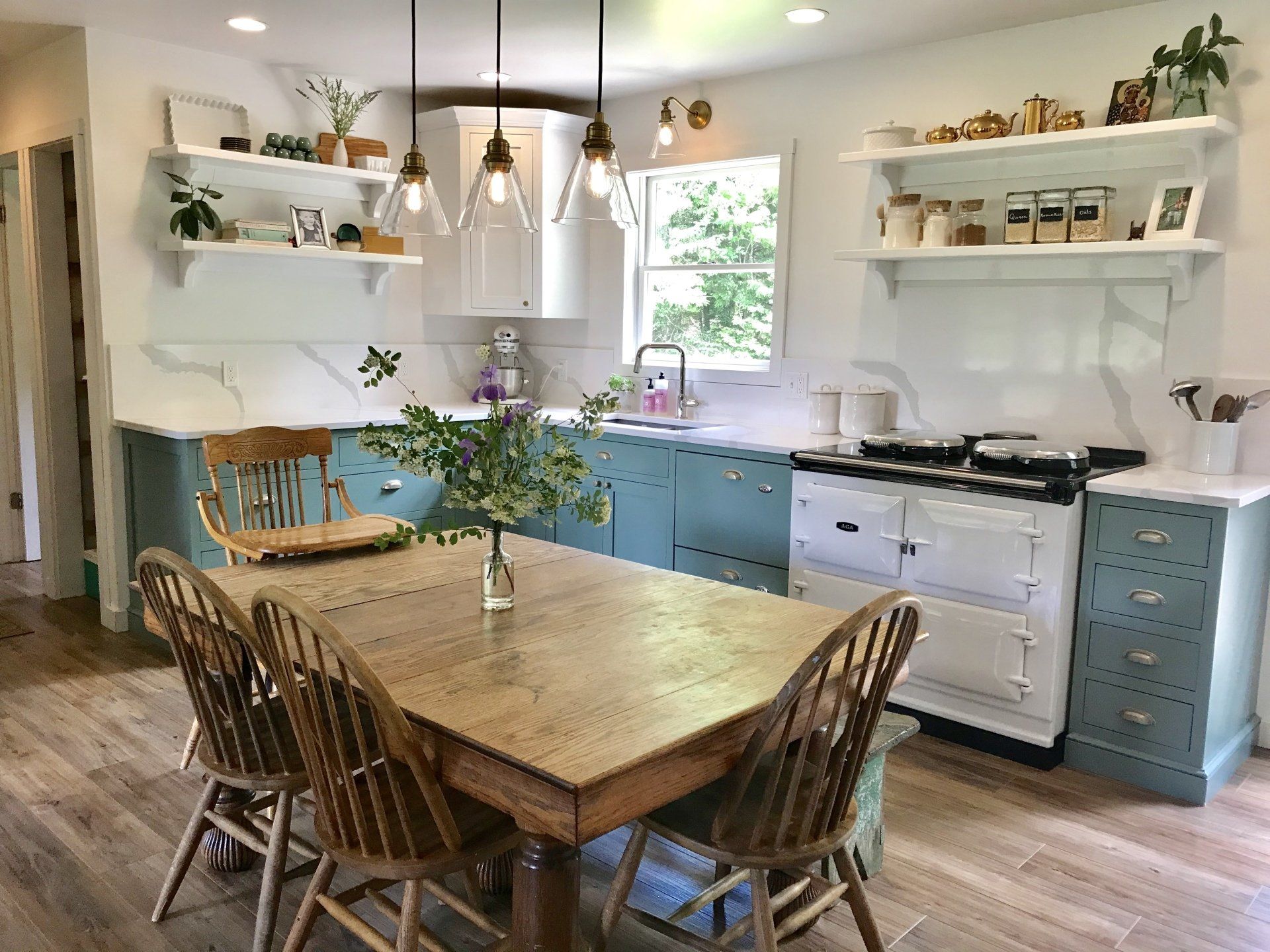 Bright kitchen with teal cabinets, white stove, wooden table, and hanging lights by a window.