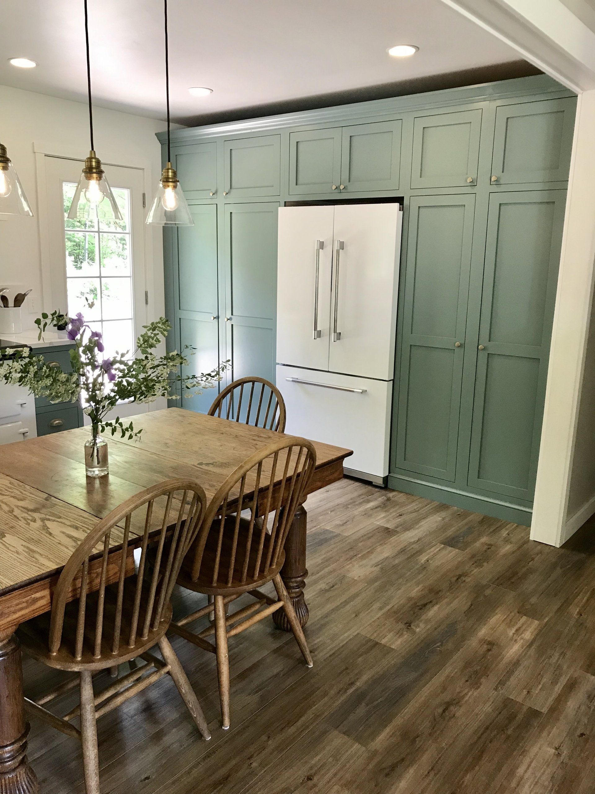 Dining area with wooden table and chairs beside a mint-green paneled wall with built-in white refrigerator cabinets.
