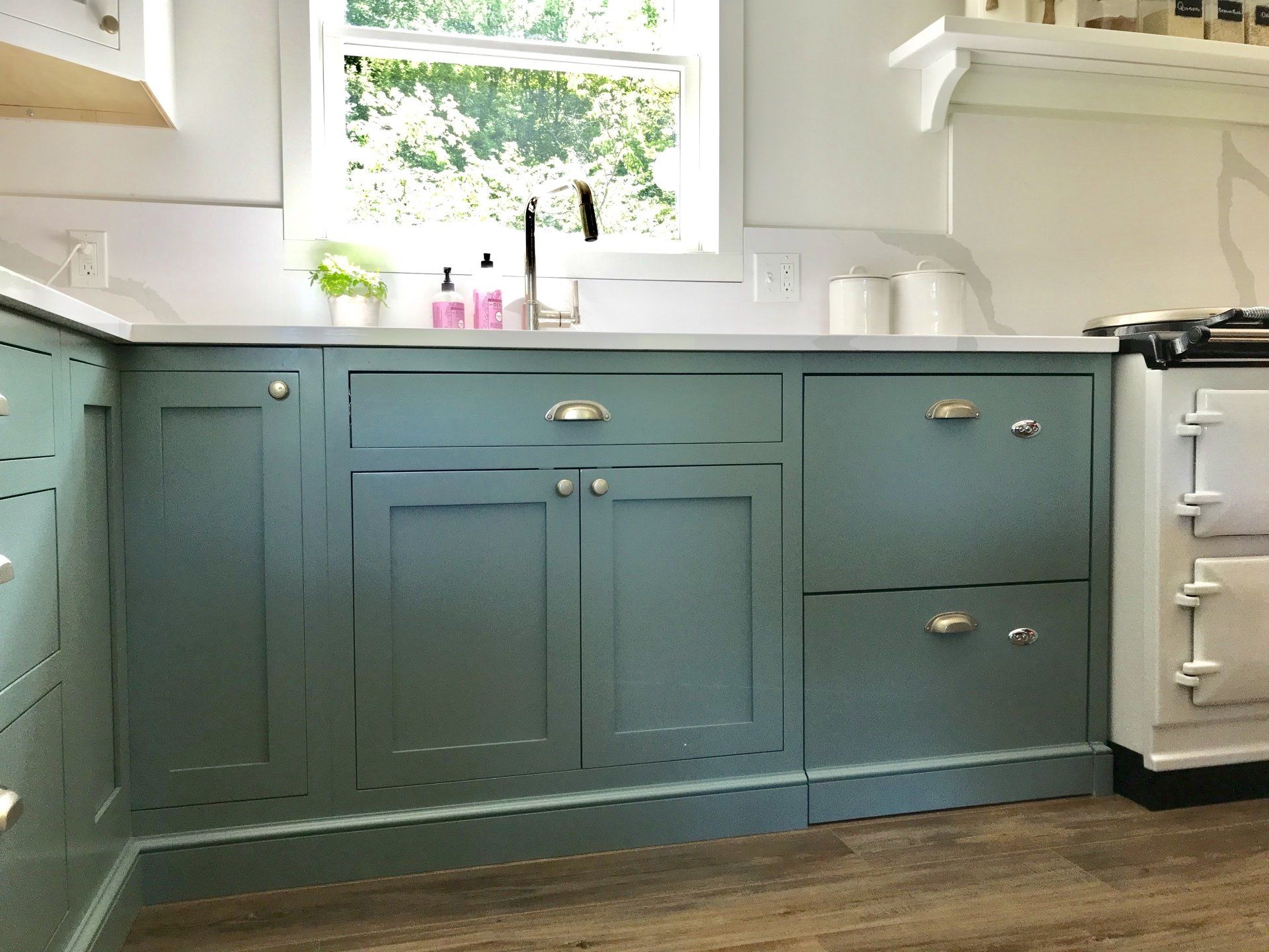 Blue kitchen cabinets under a bright window, with white countertops and a stove on the right.