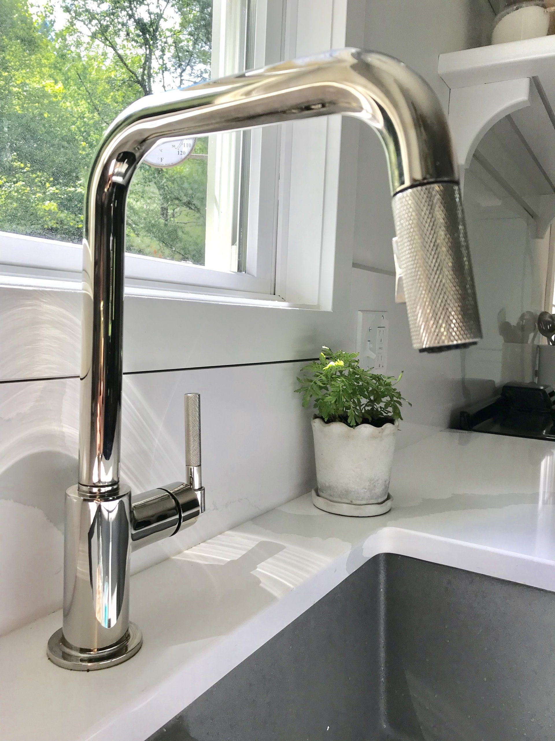 Chrome kitchen faucet over a white sink by a window with greenery outside