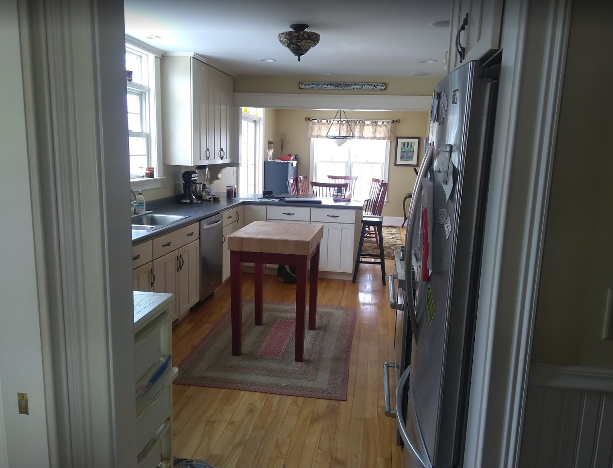 Narrow kitchen with wooden floor, central table, rug, and bright windows at the far end.