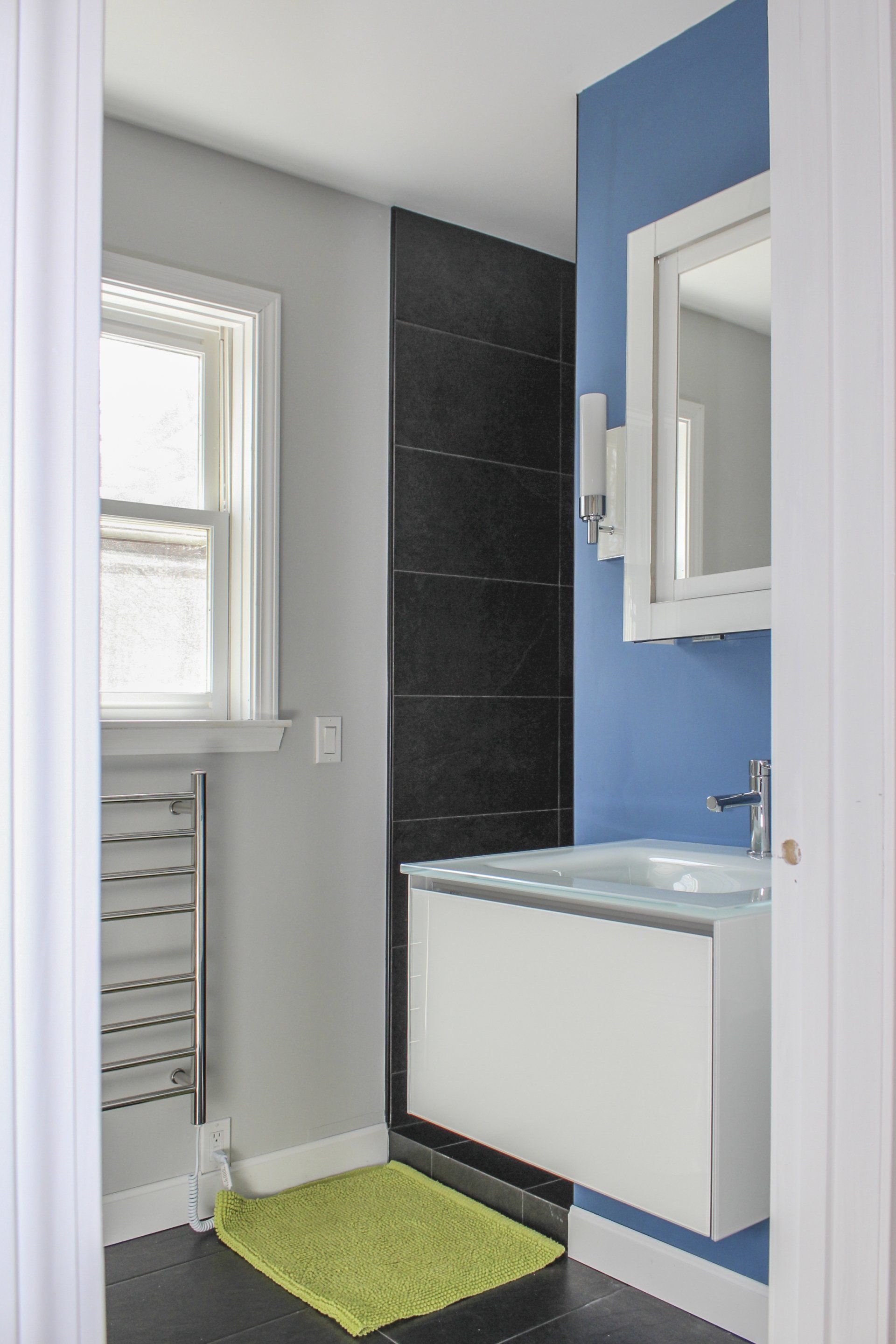 Small modern bathroom with blue accent wall, white vanity, window, and green bath mat.
