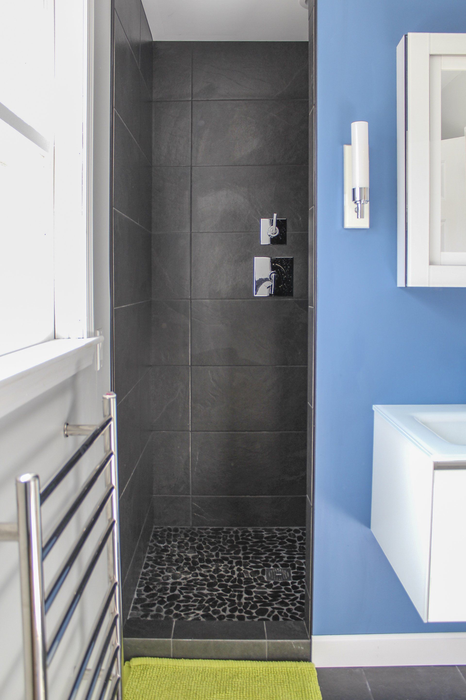 Narrow bathroom with black tiled shower, blue wall, white fixtures, and towel radiator.