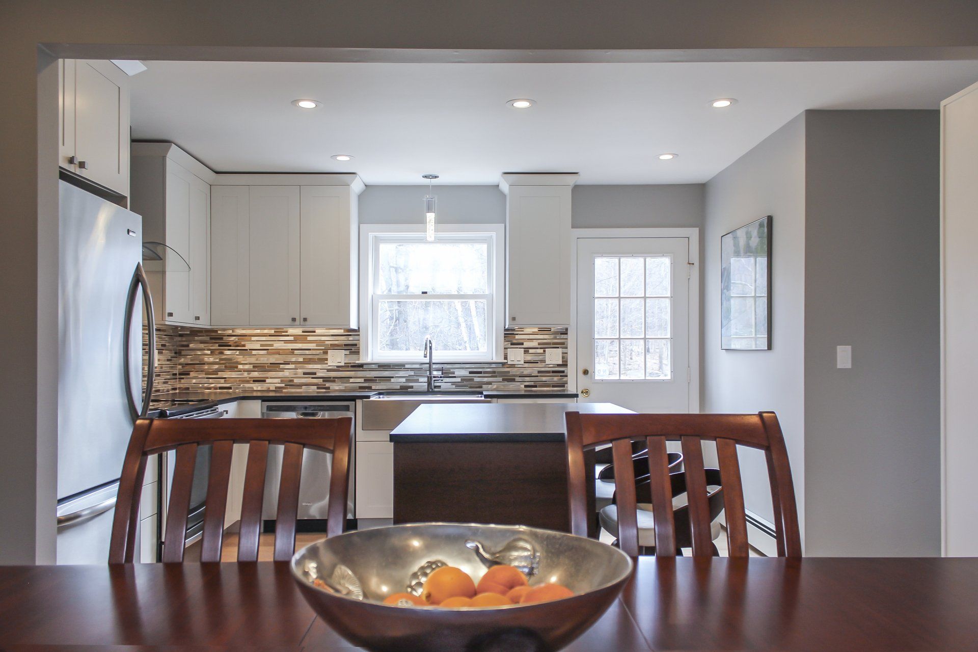 Dining table facing a bright kitchen with white cabinets, stone backsplash, and a glass door