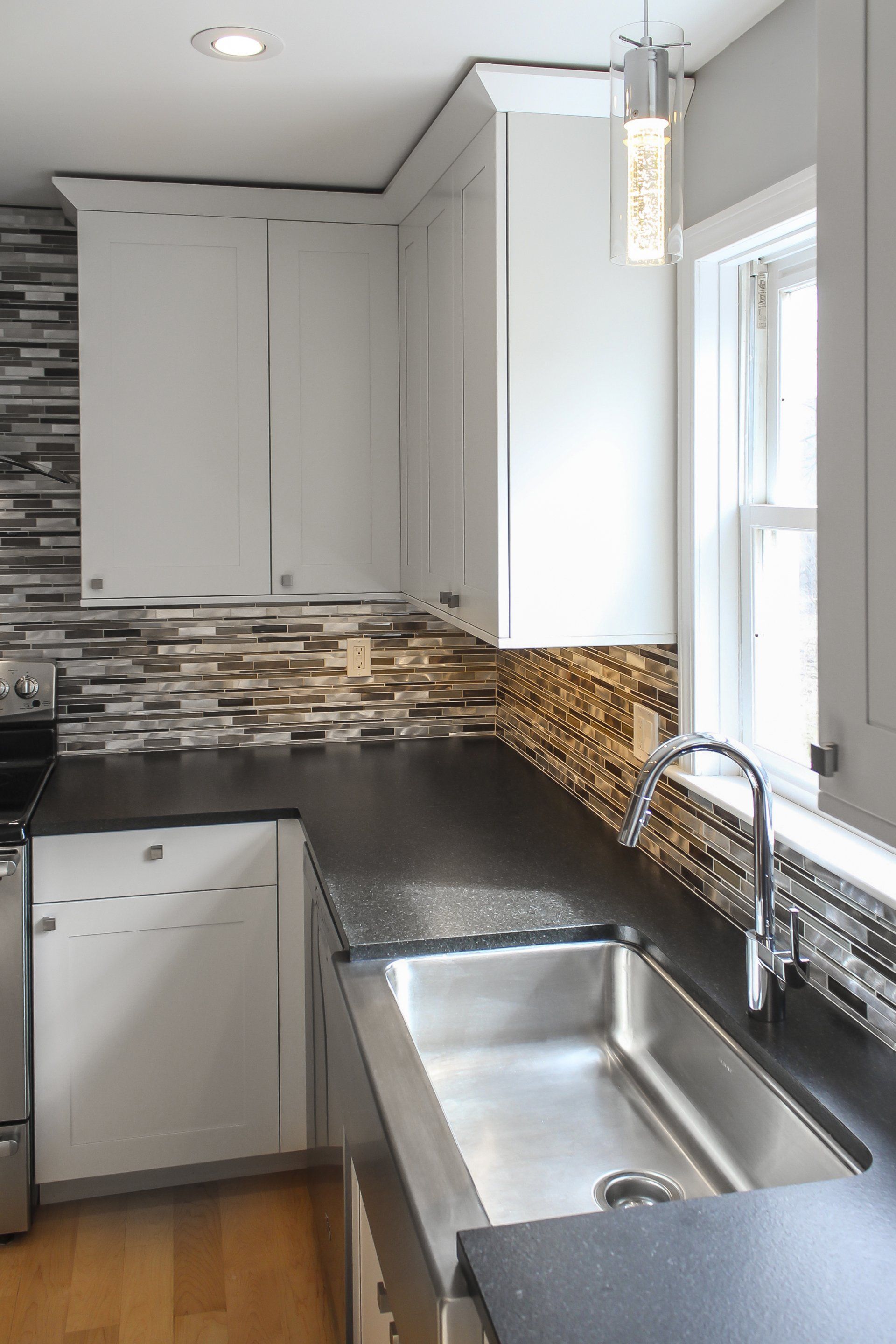 Modern kitchen corner with white cabinets, black countertop, mosaic backsplash, and stainless-steel sink by a window
