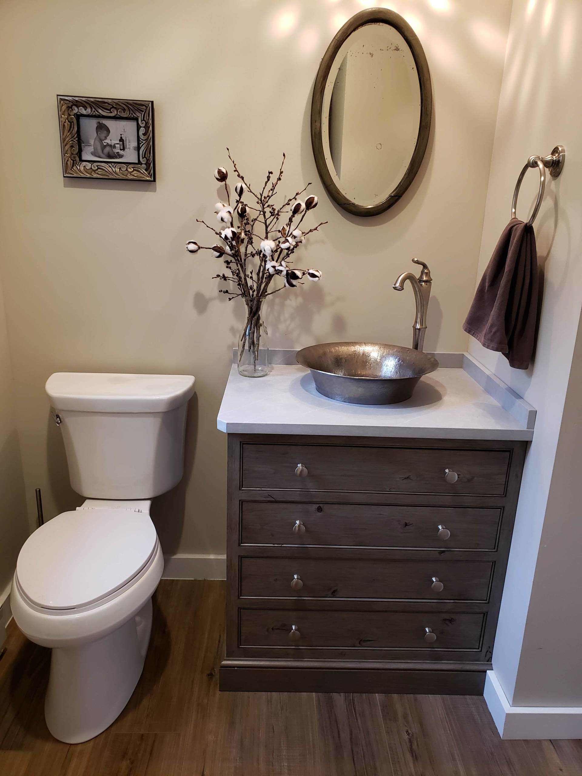 Small bathroom vanity with oval mirror, dark wood cabinet, white countertop, wall decor, and a toilet beside it
