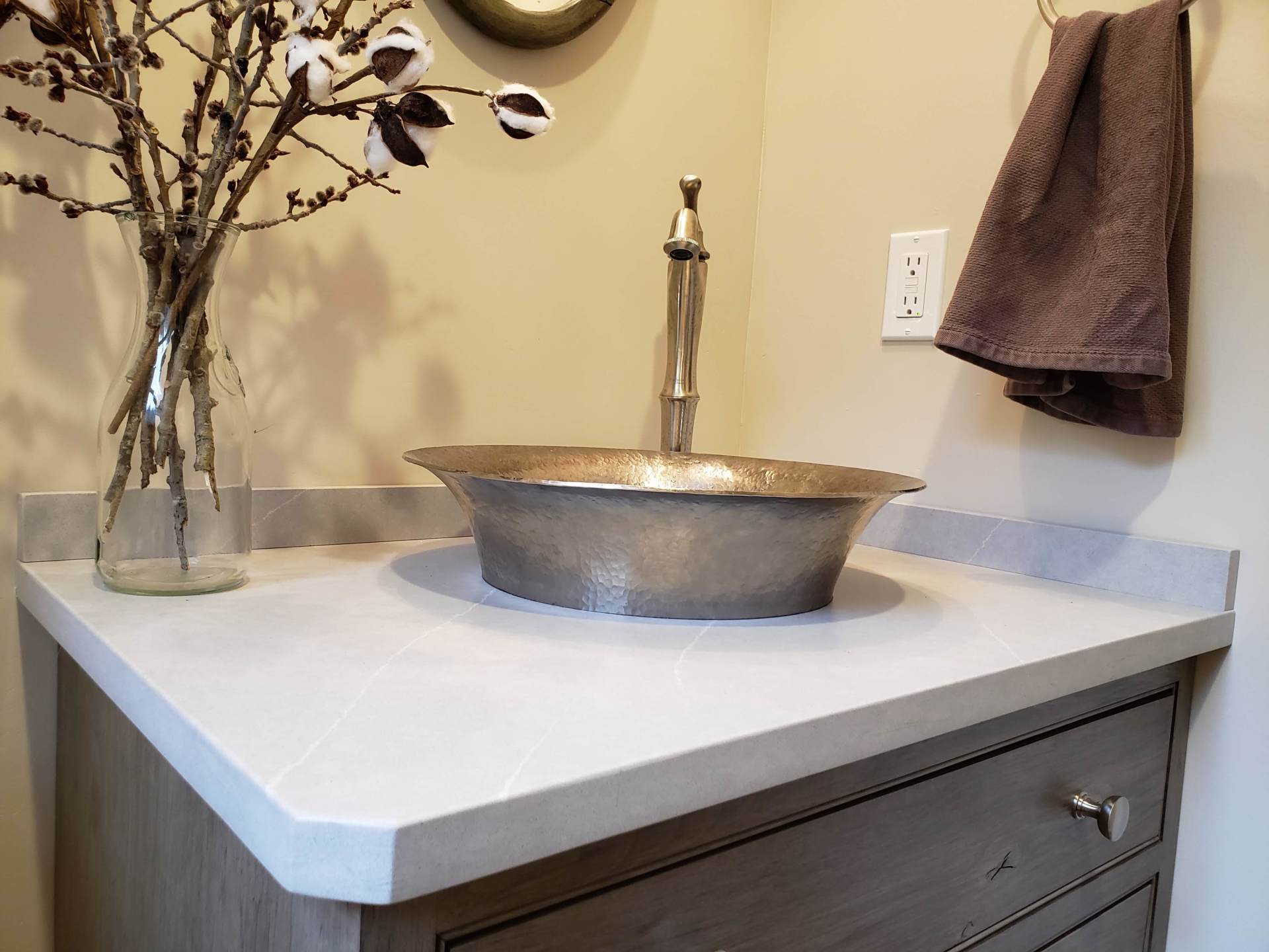 Bathroom vanity with a vessel sink, faucet, wall towel, and vase of branches on a light countertop