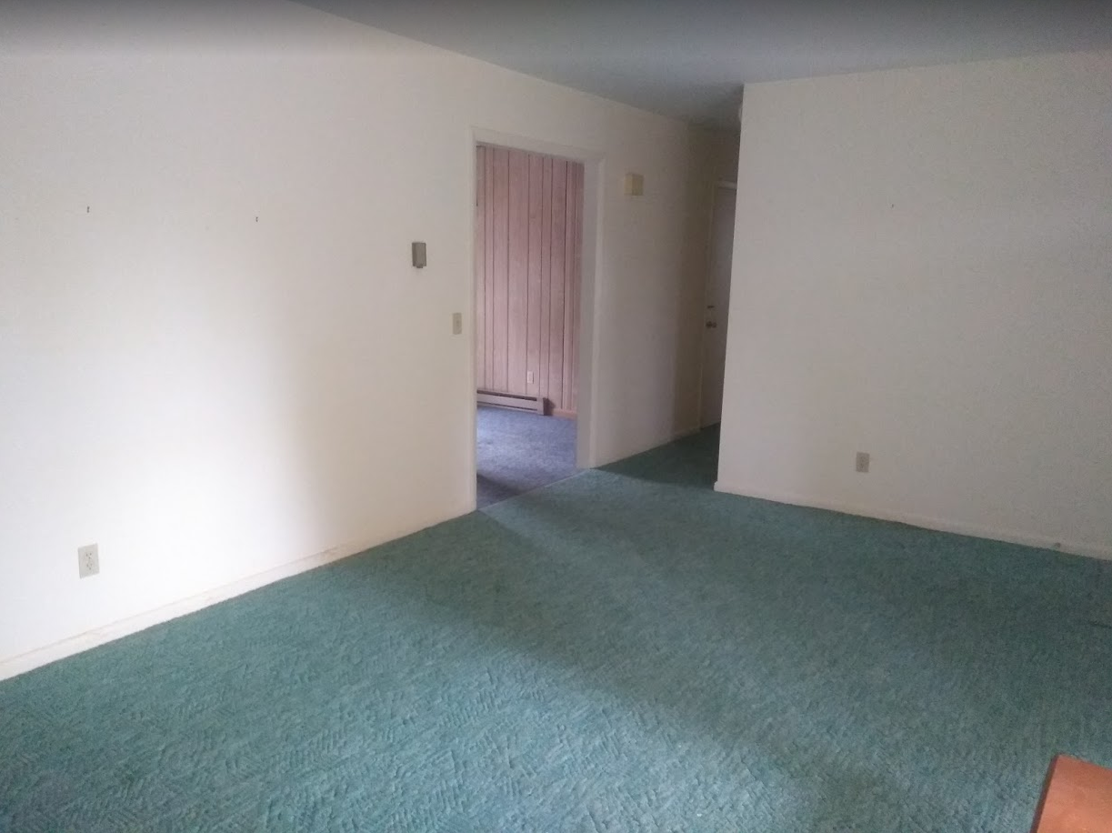 Empty carpeted living room with white walls and an open doorway to a hallway.