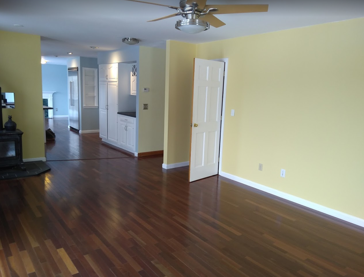 Empty living room with dark wood floors, yellow walls, and ceiling fan, leading into a hallway.