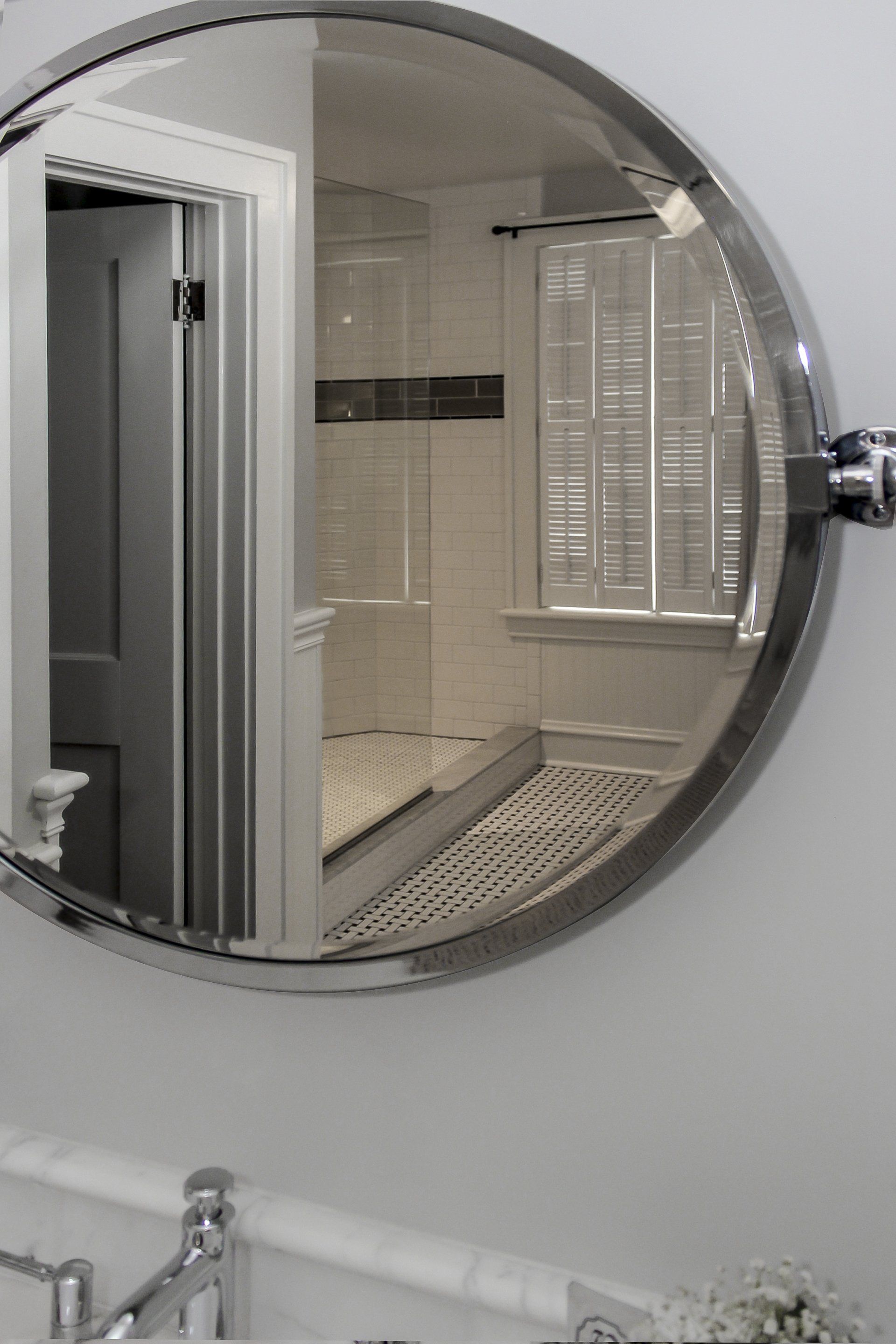 Round bathroom mirror reflecting a white tiled shower and window with shutters