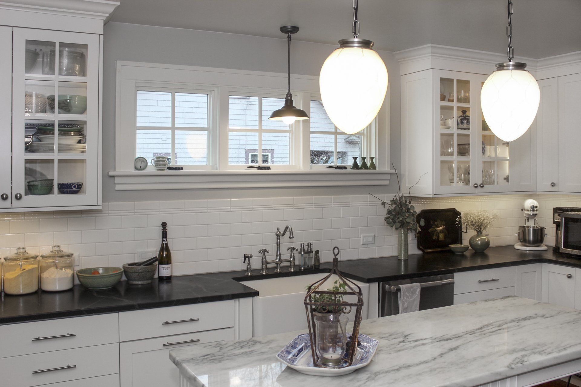 Bright white kitchen with marble island, black countertops, glass cabinets, and hanging pendant lights.