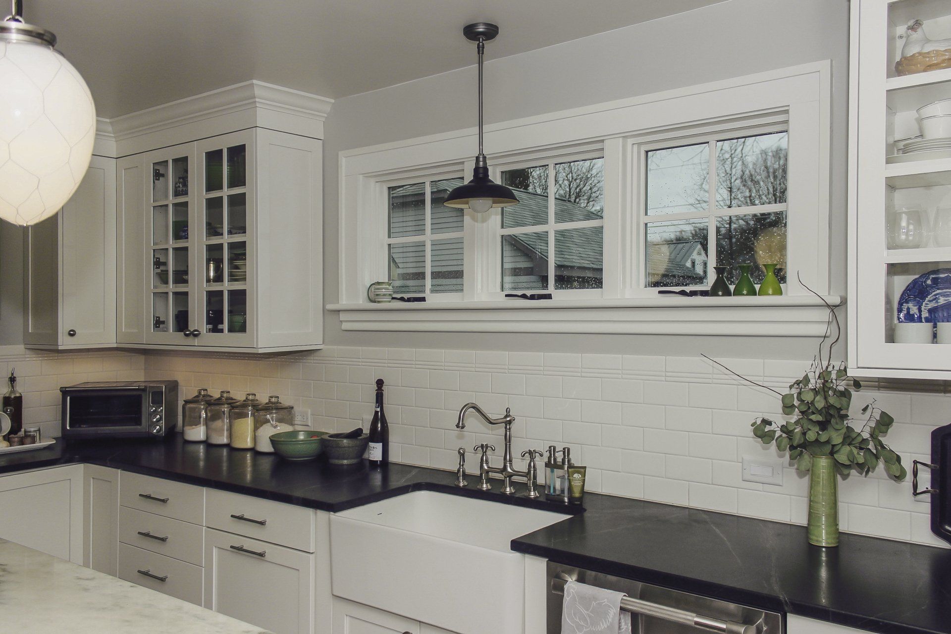 Modern white kitchen with black countertops, farmhouse sink, window, and pendant light
