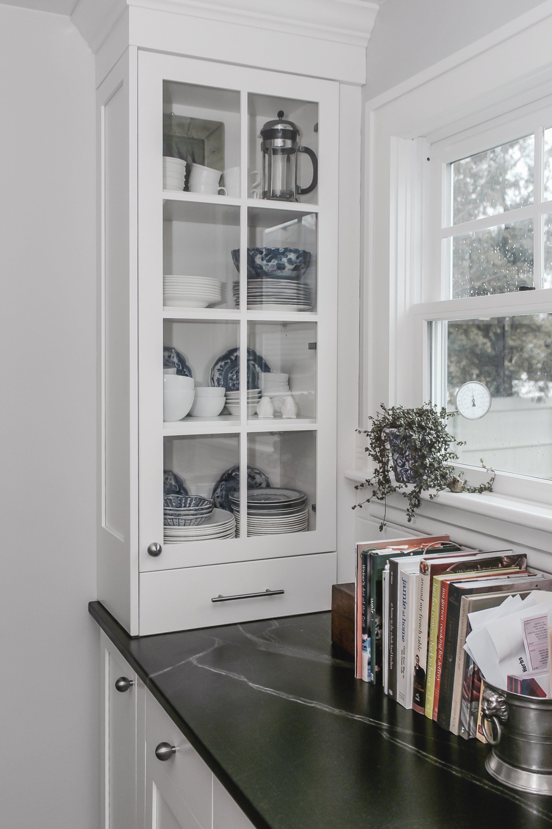 White kitchen corner with glass cabinets, dishes, and a window above a dark countertop lined with magazines.