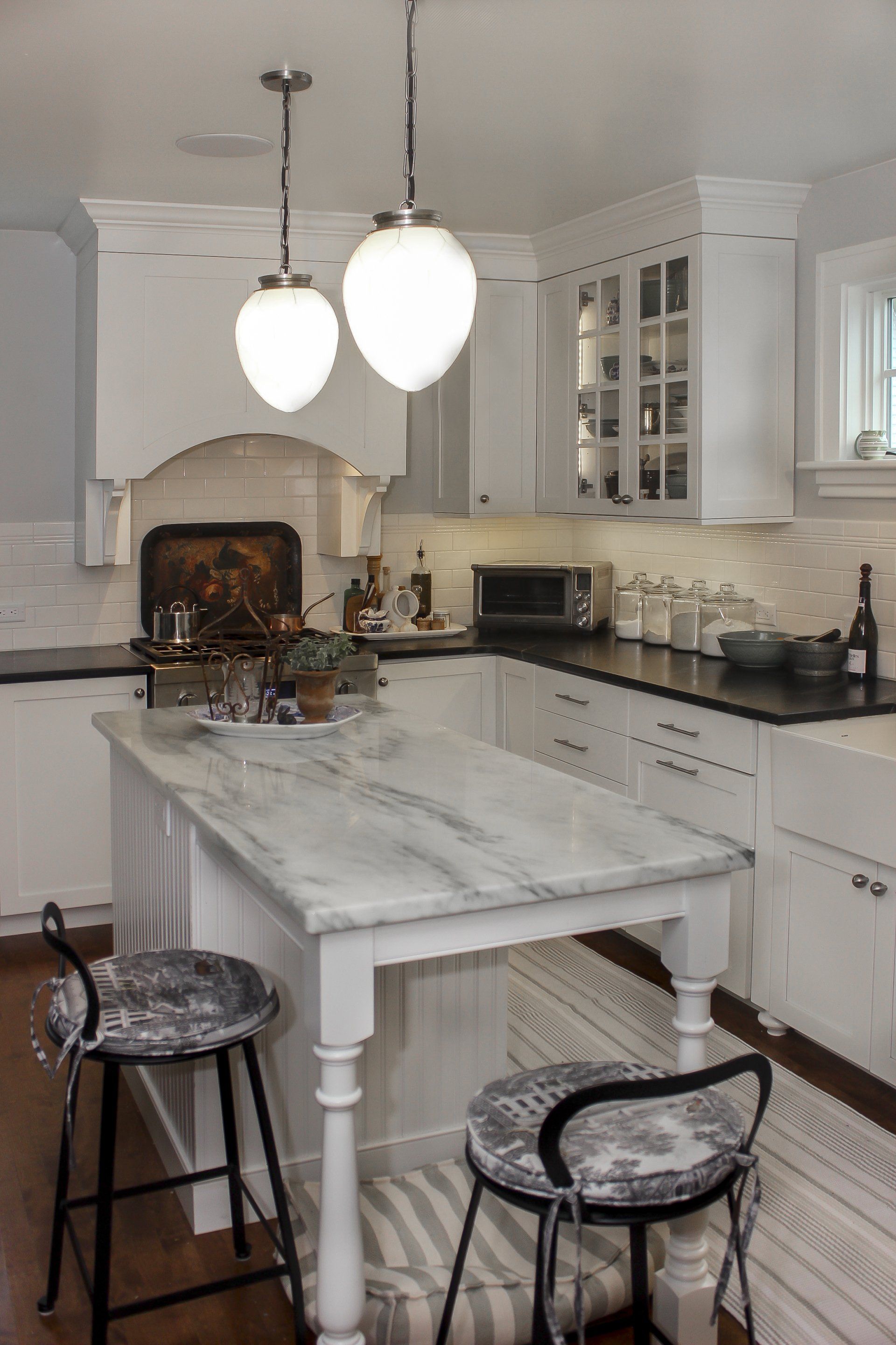 White kitchen with marble island, pendant lights, black stools, and a stove fireplace in the background