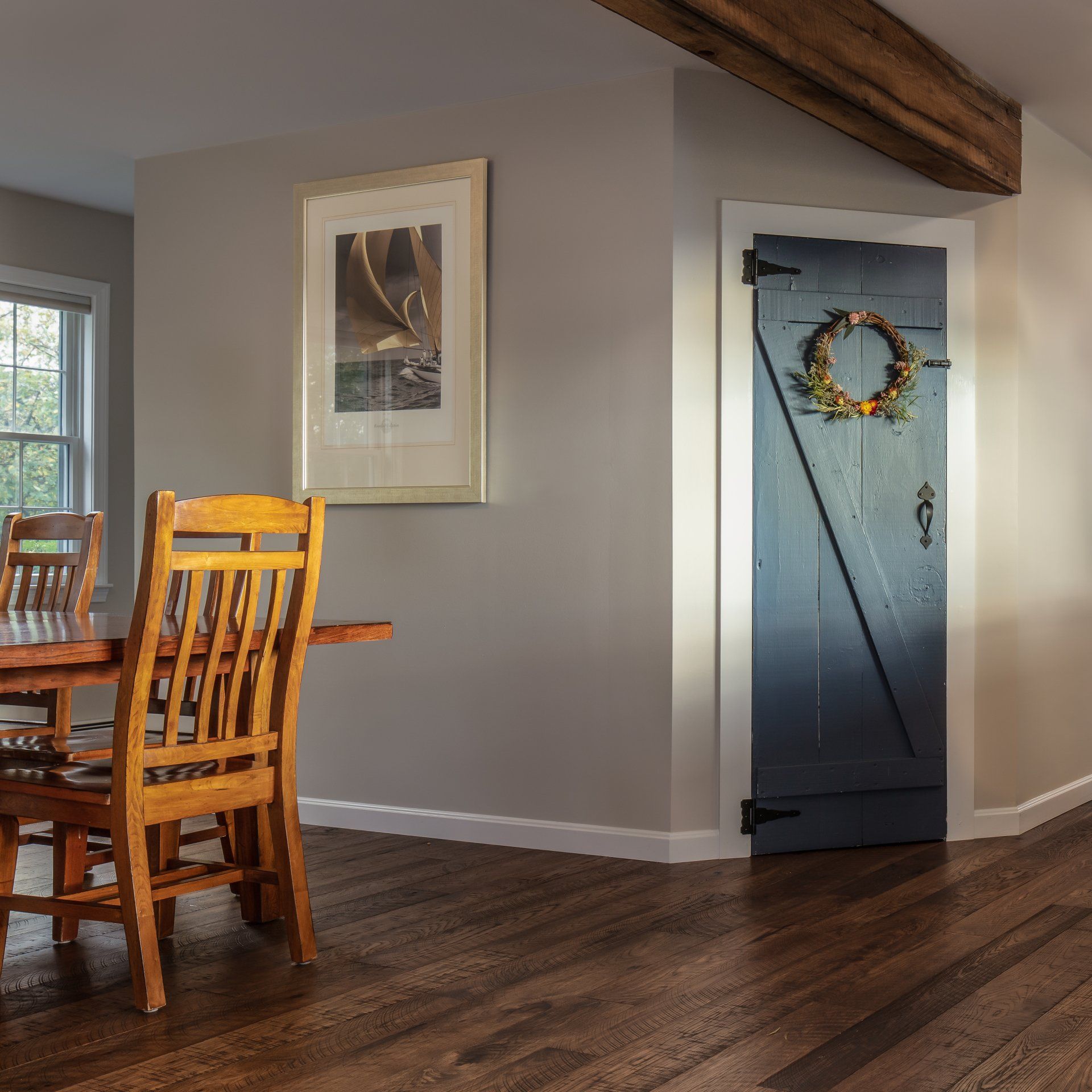Dining area with wooden table and chairs, blue door with wreath, and framed art on gray walls.