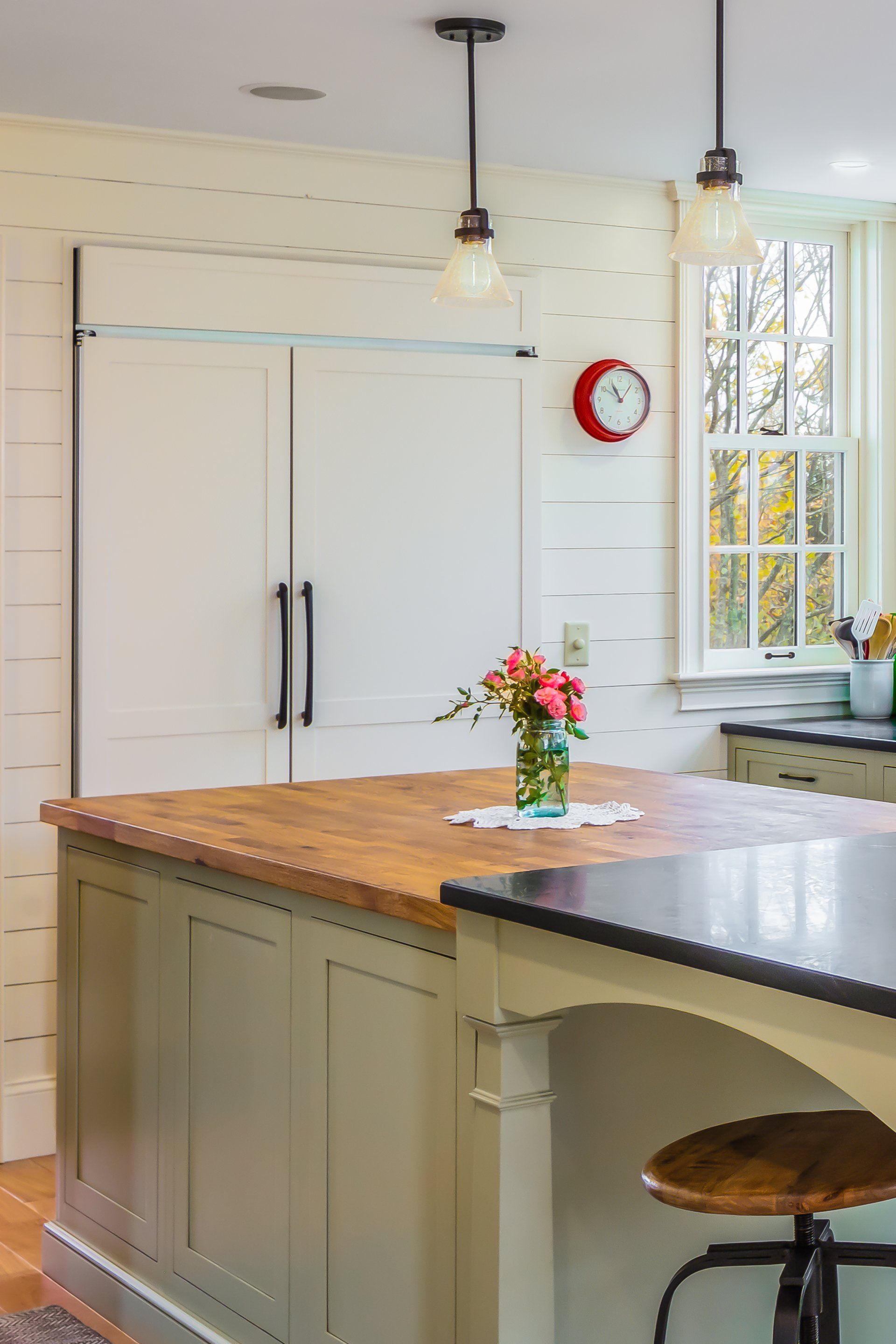 Bright kitchen with white cabinets, wood island, hanging lights, red wall clock, and a flower centerpiece.