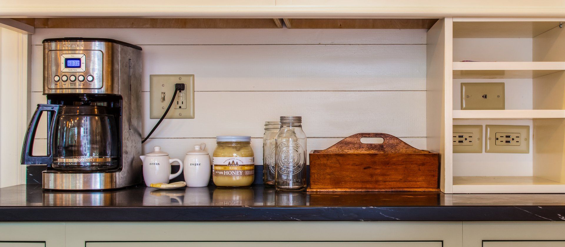 Kitchen counter with coffee maker, cups, jars, wicker basket, and wall outlet under shelves