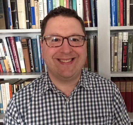 Robert M. Mello, Jr., LICSW, MSW, MA, CSAT, EMDR II Member, New England Recovery Associates | Serving Westborough, South/Central Massachusetts, Rhode Island, Northern Connecticut A smiling person with glasses wearing a checkered shirt, positioned in front of a full bookshelf.