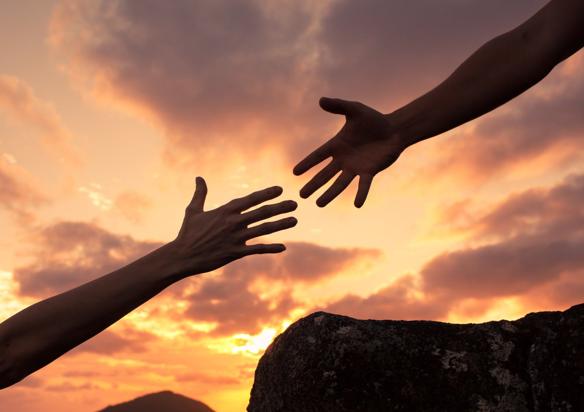 Two hands reaching toward each other against a bright, orange sunset, with one hand stretching from a rocky ledge.