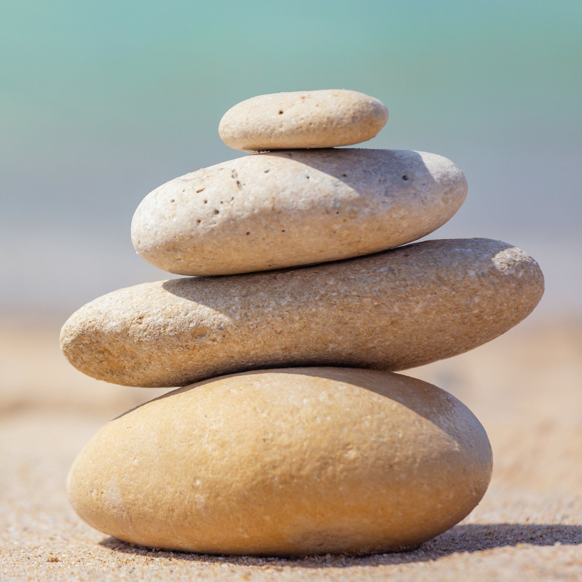 A stack of four smooth, light-colored stones balanced on top of each other on a sandy beach with the ocean in the background.