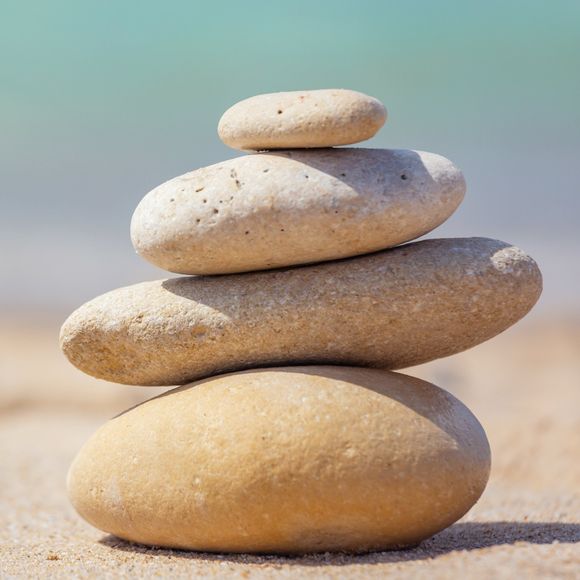 A stack of four smooth, light-colored stones balanced on top of each other on a sandy beach with the ocean in the background.