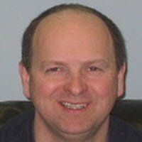 A smiling person with a receding hairline, wearing a dark shirt against a light gray background.