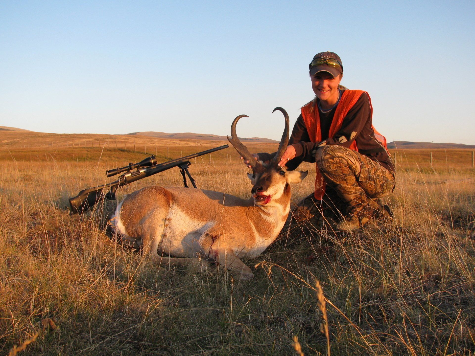 Hunter kneeling next to a pronghorn antelope and rifle in a field at sunset.