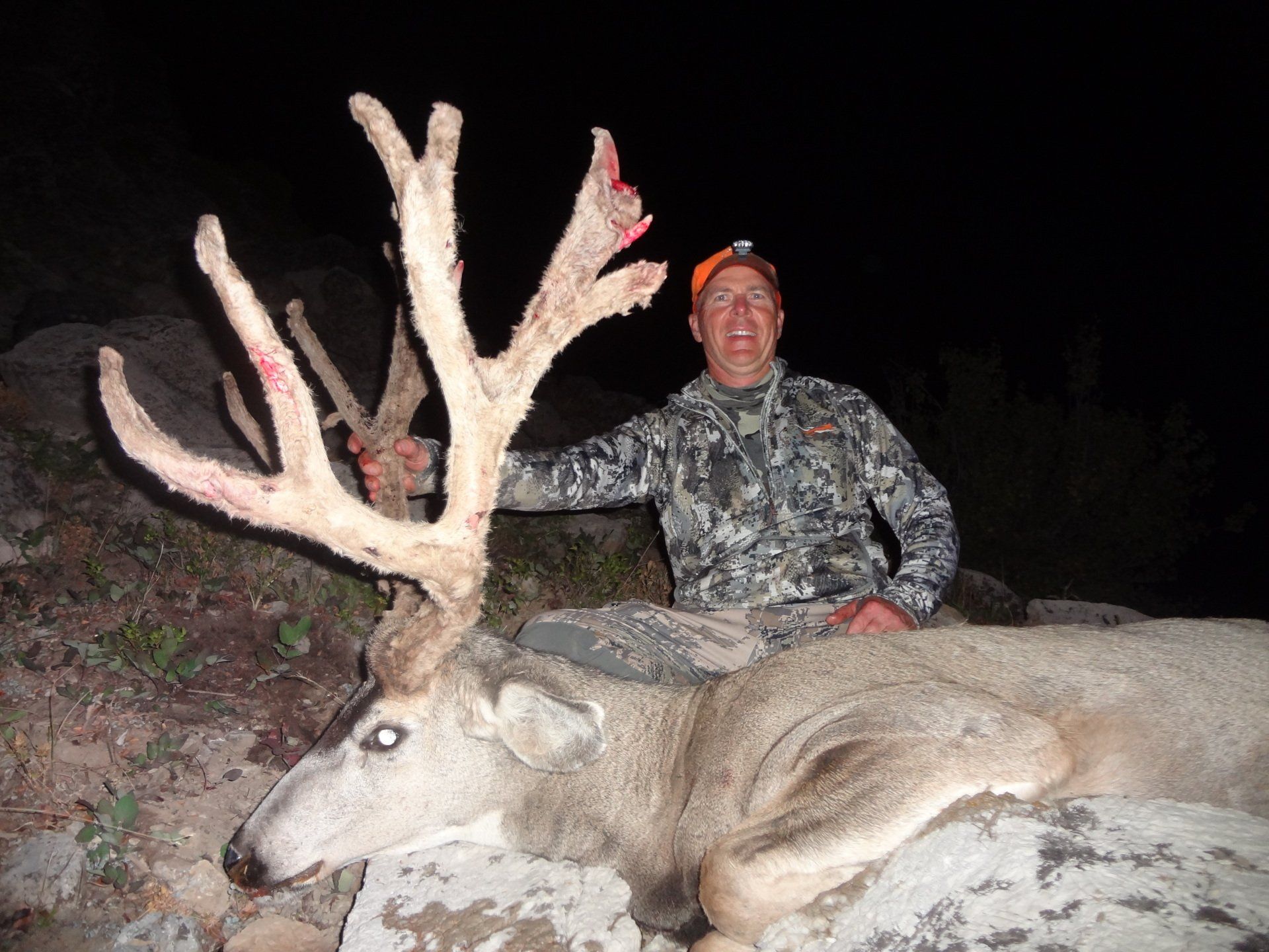 Hunter kneels beside a large buck with massive antlers, night setting. Hunter wears camouflage and a headlamp.