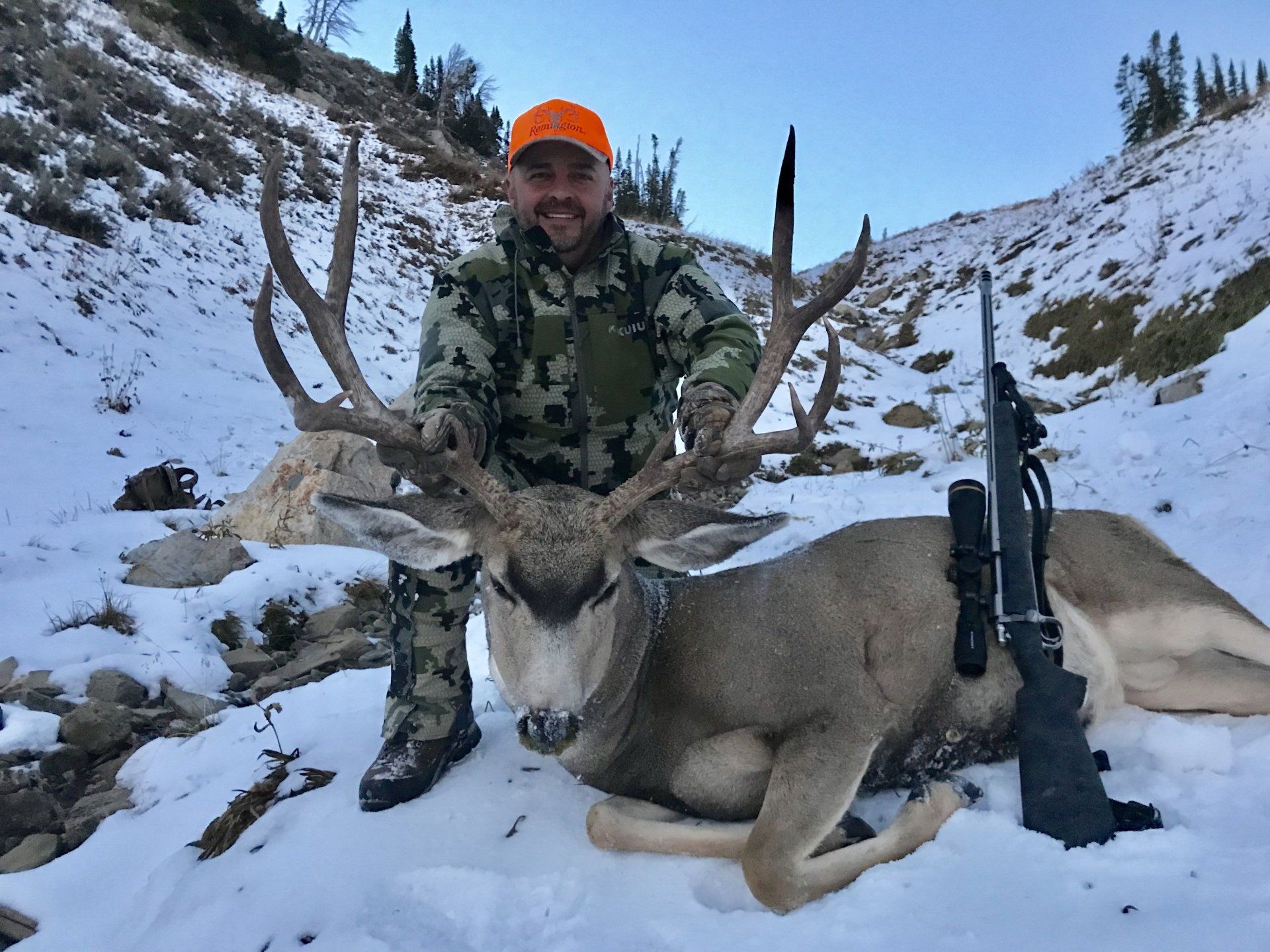 Hunter in camouflage with a large buck in the snow; rifle nearby.