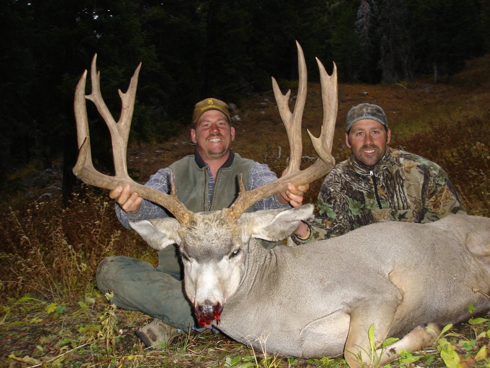 Two men holding large deer antlers in a wooded area, deer on ground with bloody snout.