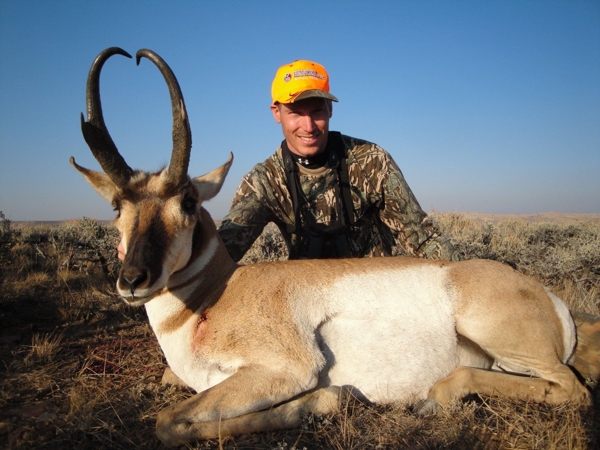 Man in camouflage with a pronghorn antelope on the ground; outdoor setting, blue sky.