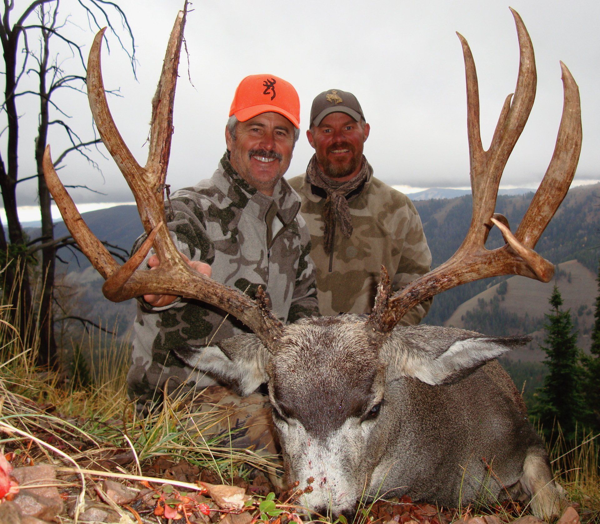 Two men pose with a large buck with antlers. They are outdoors, smiling, dressed in camouflage.