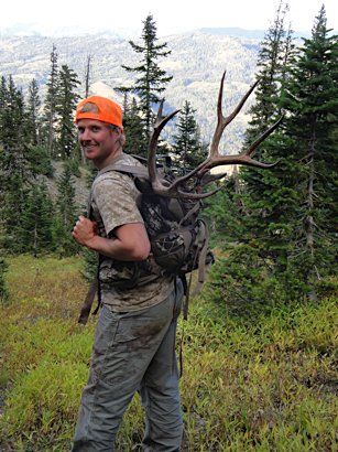 Man with elk antlers on backpack in a forest, wearing orange hat and camo clothing, smiling.