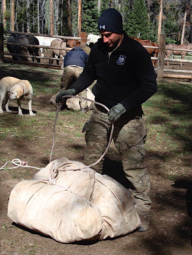 Man in camo pants and black jacket ties rope around two large canvas bags outdoors.