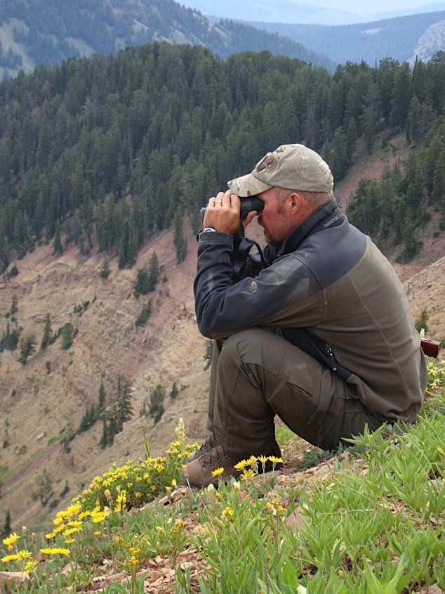 Man seated, using binoculars, overlooking a forested mountainside, green vegetation in foreground.