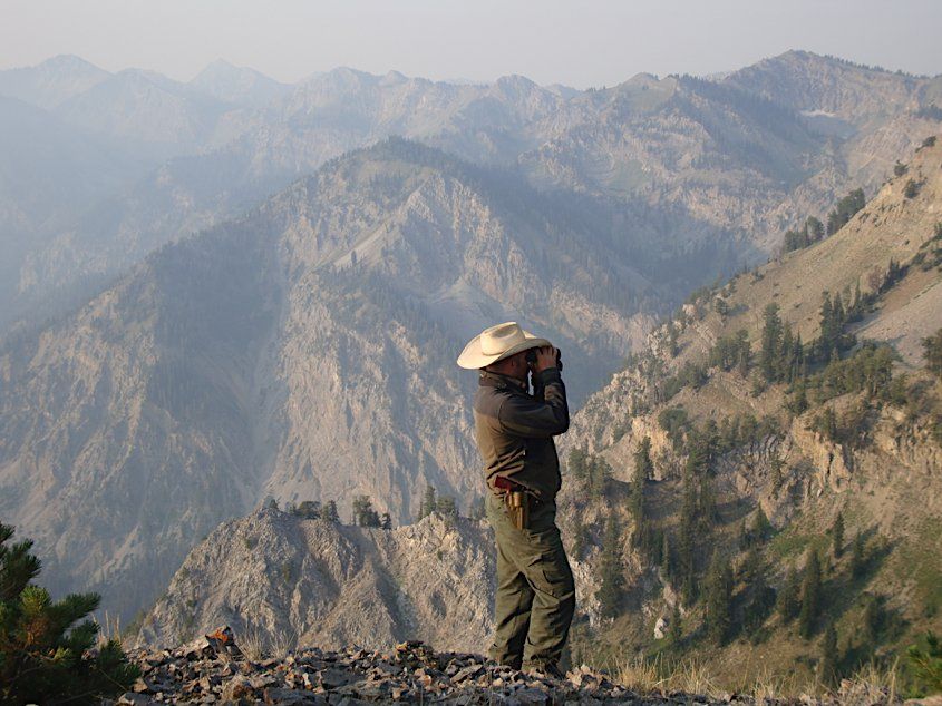 Person with a hat photographs a mountain range, standing on a rocky ledge.