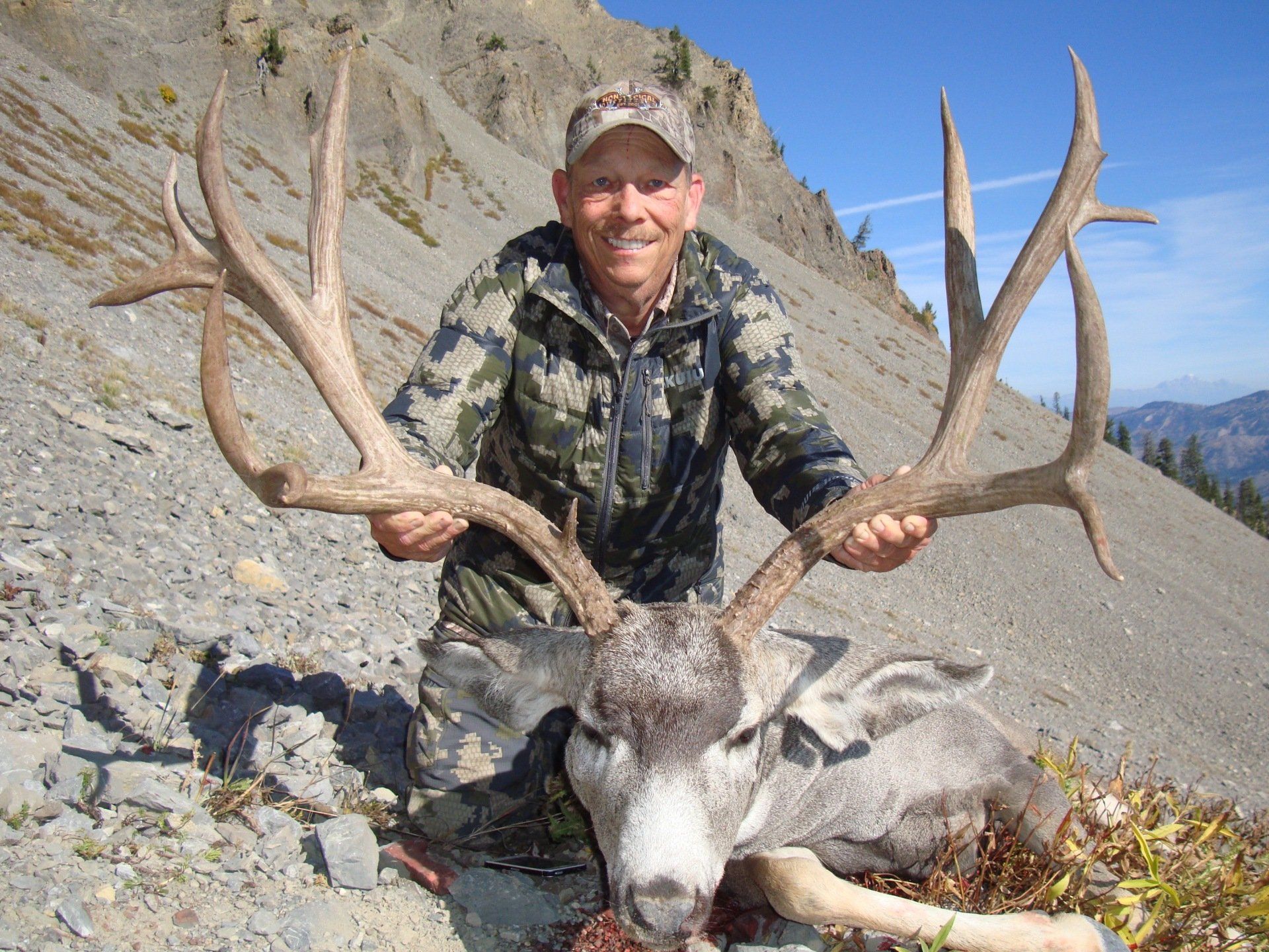 Hunter kneeling, holding large mule deer antlers. Mountain backdrop, camouflage clothing, smiling.