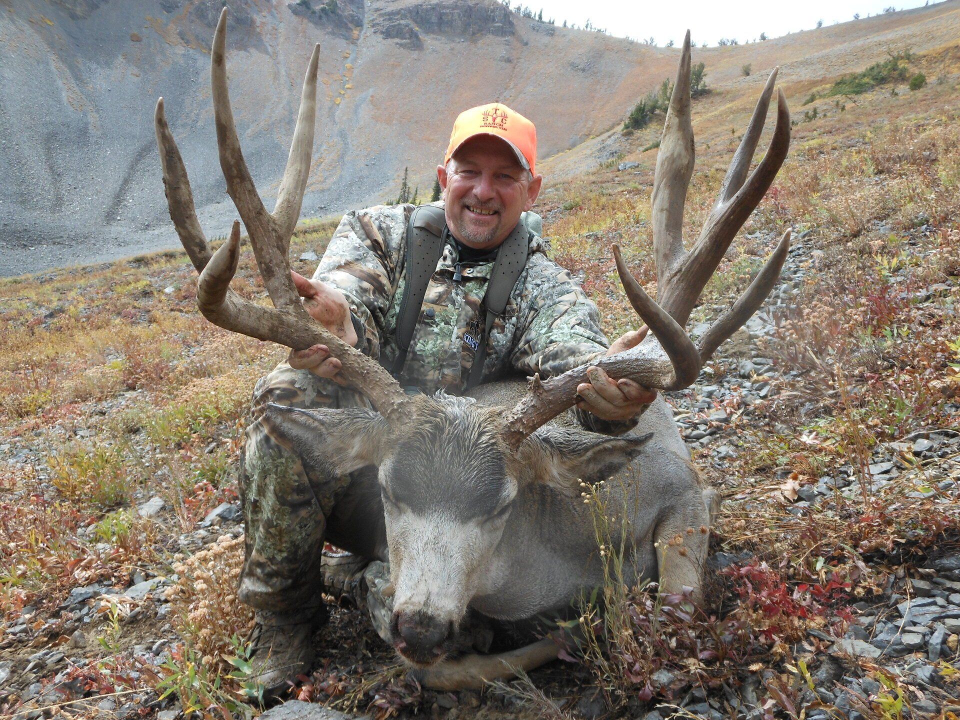 Hunter in camo kneeling next to a large deer with impressive antlers, orange hat, outdoors.