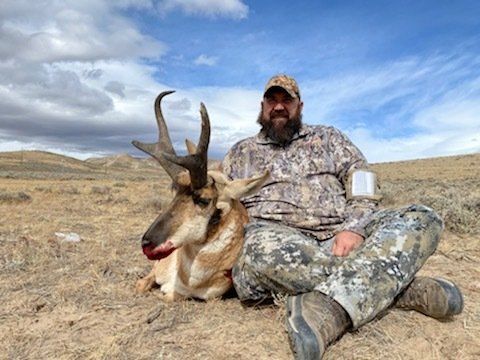 Hunter sits next to a trophy harvested pronghorn antelope in a field. Cloudy sky.