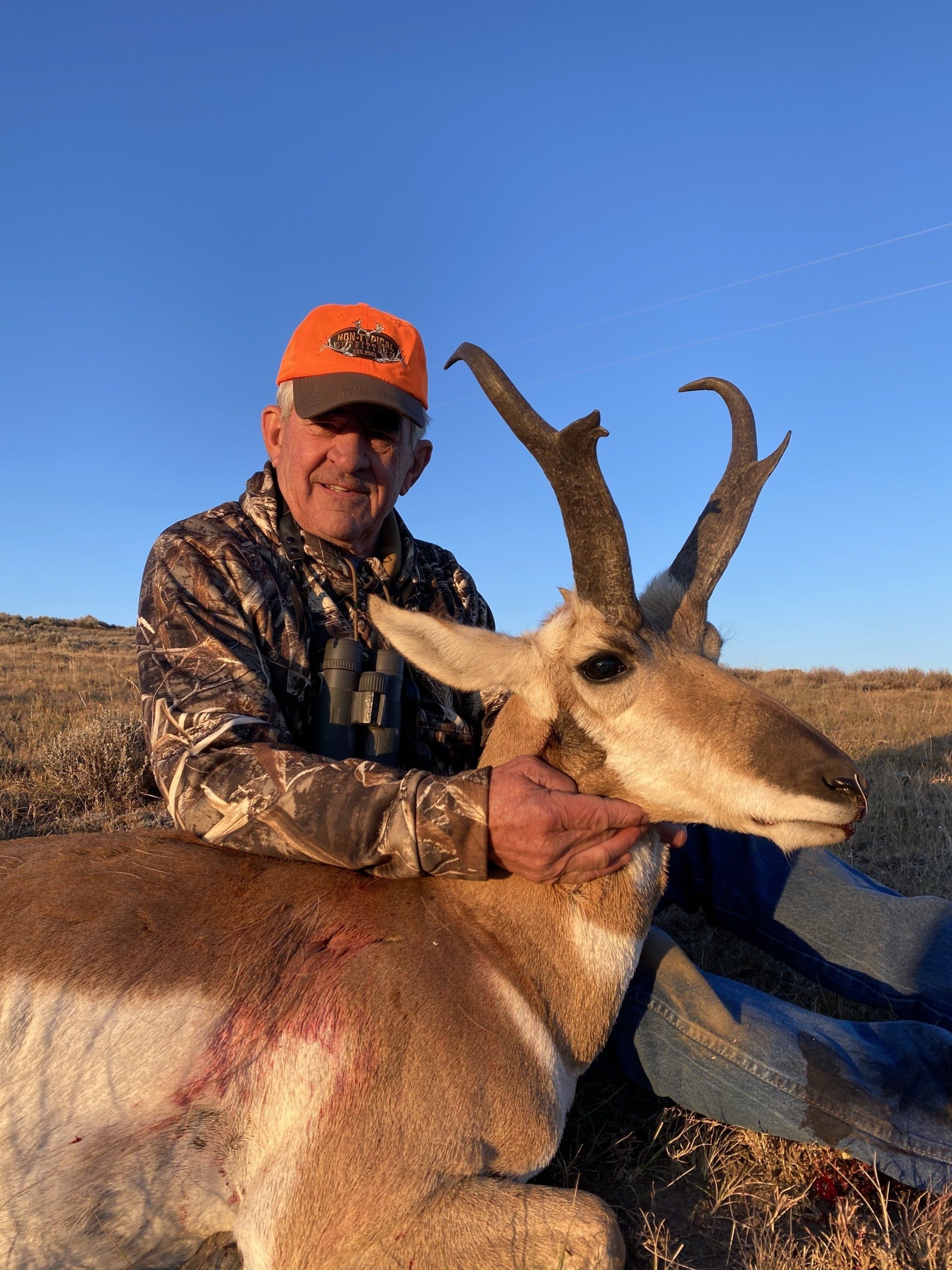 Hunter in camouflage holds a pronghorn antelope, blue sky background.