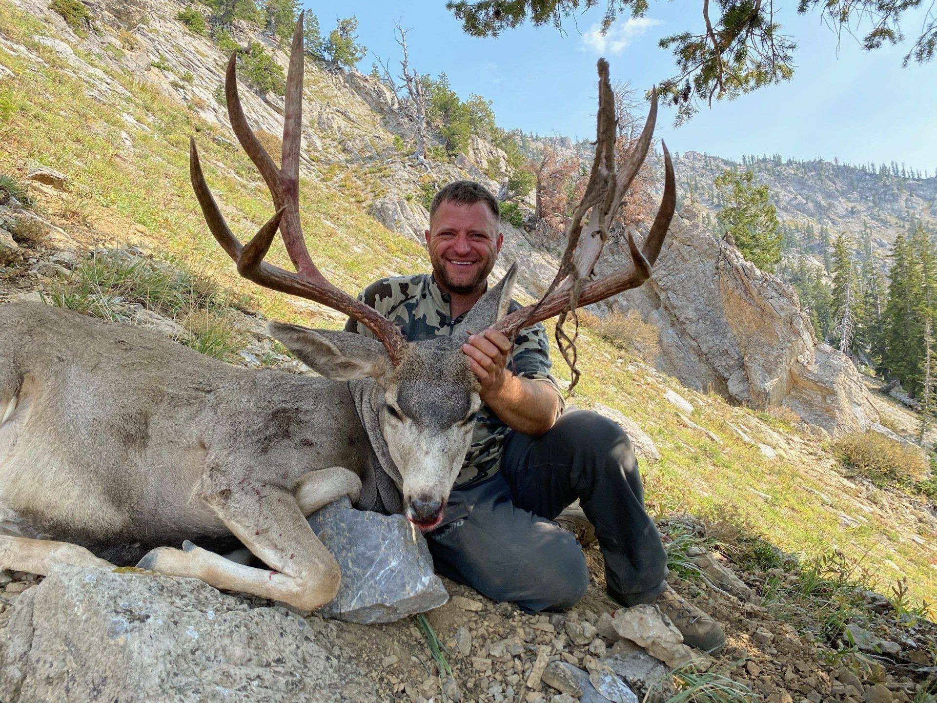 Man kneels beside a large dead deer, holding its antlers. Mountainous, rocky terrain in background.