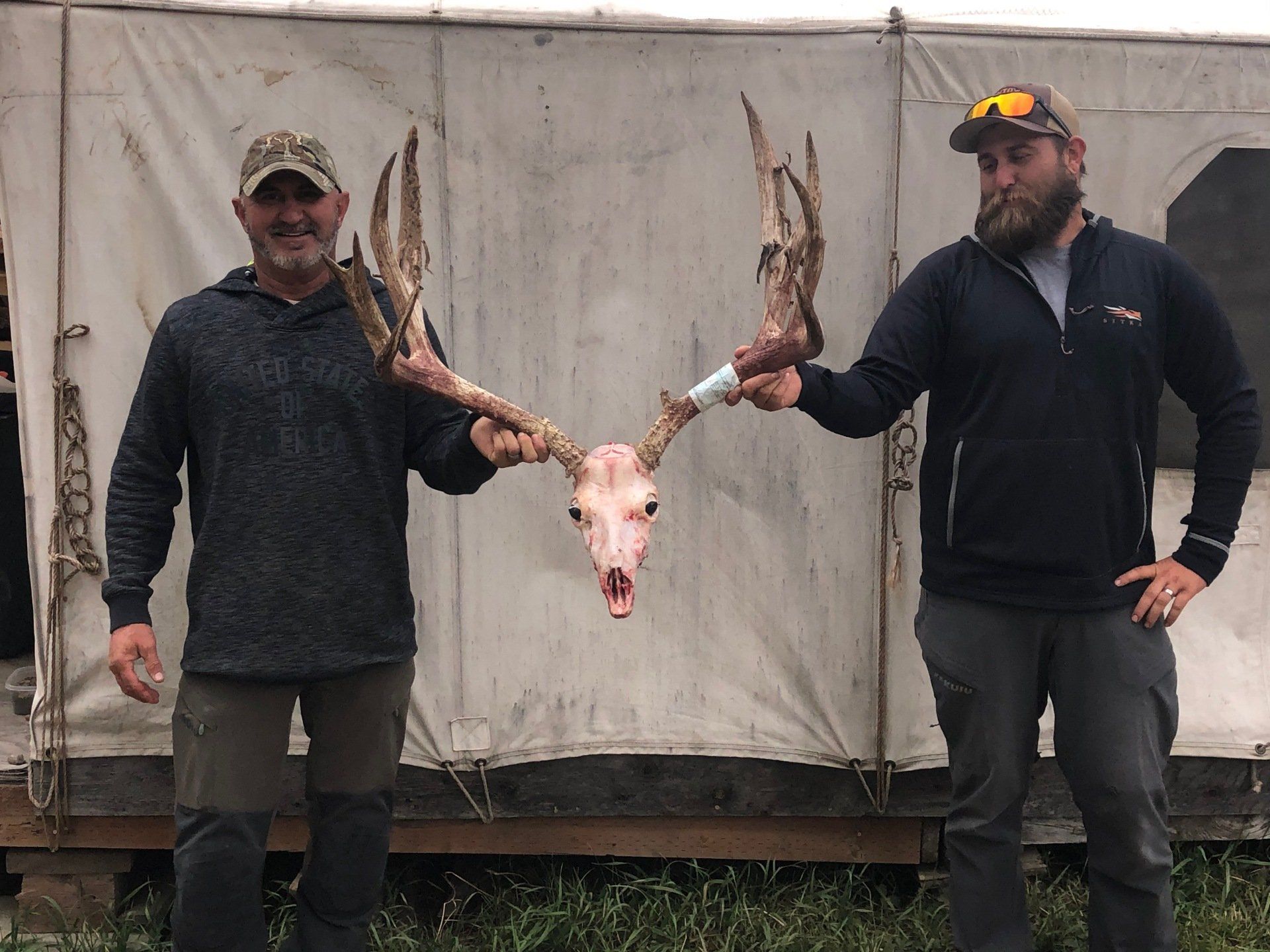 Two men hold a deer skull with large antlers in front of a tent. They smile, one wearing a camo hat.