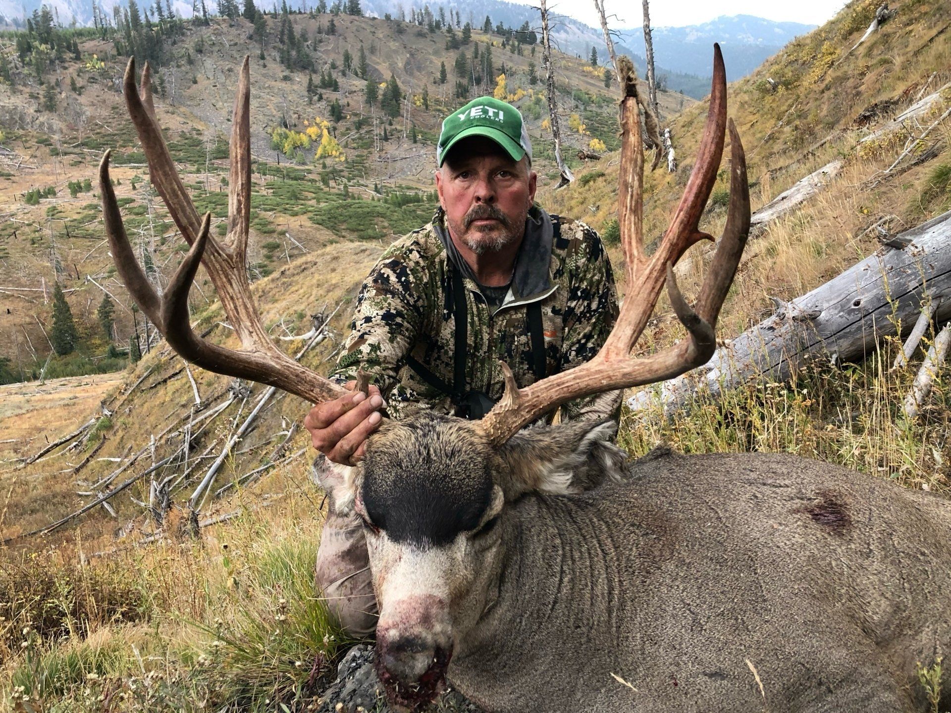 Man in camo holding large buck with impressive antlers in a mountain setting.