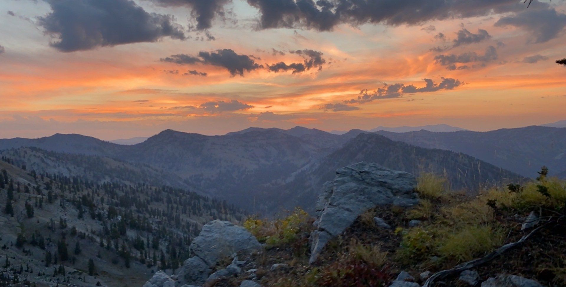 Sunset over mountain range; orange and yellow sky, silhouetted peaks, rocky foreground.