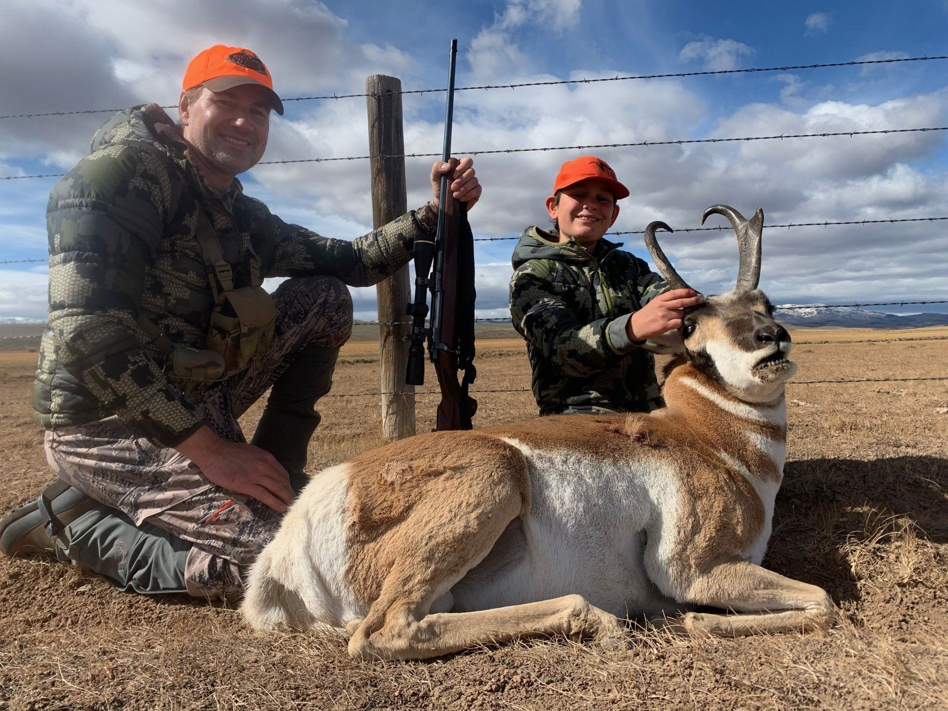 Father and child posing with a harvested pronghorn antelope in a field, both wearing camo.